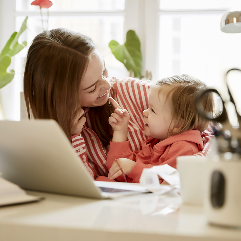 Leende mamma sitter med flicka vid skrivbord på hemmakontor